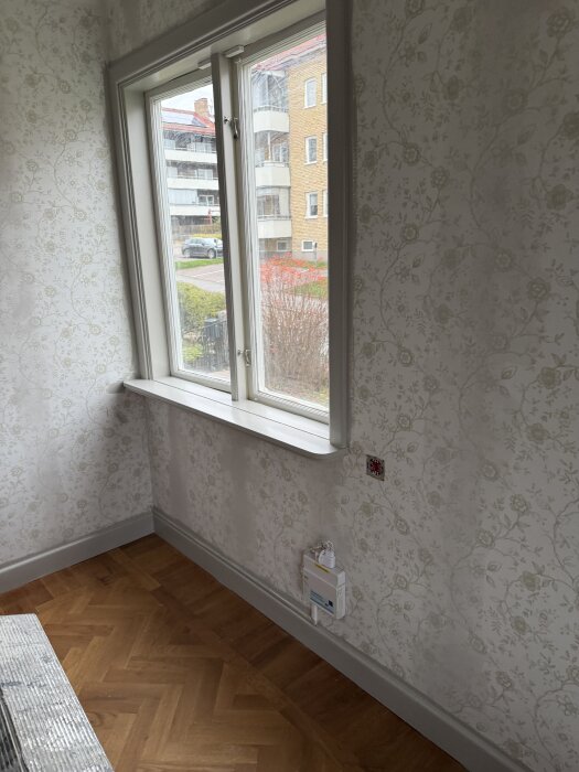 Fönster inomhus med blommiga tapeter och parkettgolv, vy mot gata utanför.