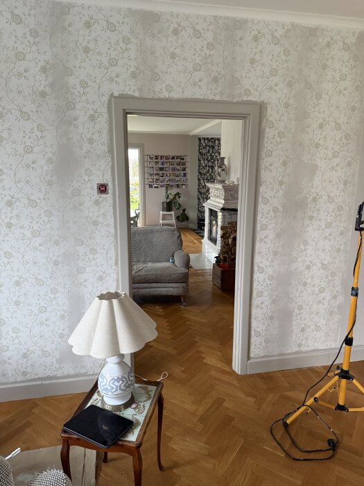 Öppning mellan renoverat TV-rum och vardagsrum, med blommig tapet, vit lampa på sidobord, och del av orange bygglampa synlig.