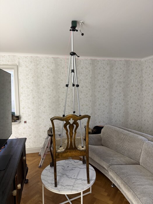 Trebent stativ med laser på stol, ovanpå marmorbord i renoveringsrum med blommig tapet och grå soffa.