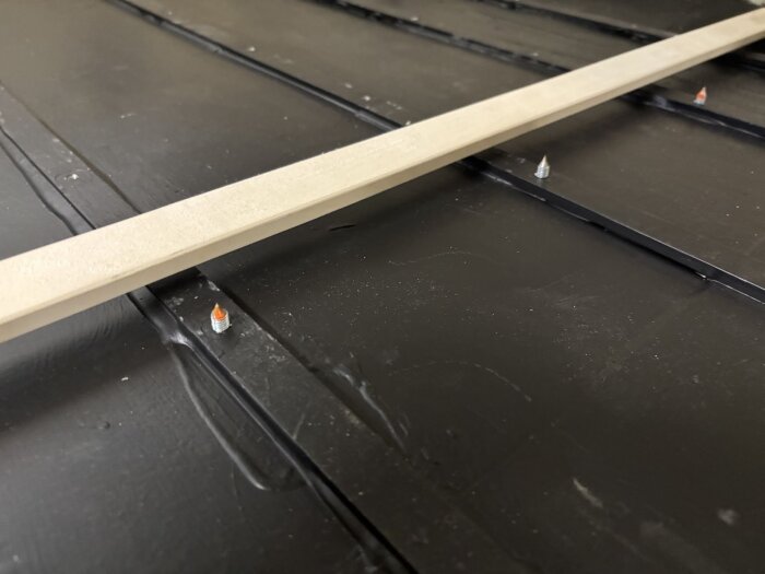 Träspjäla av asp placerad på svart underlag med synliga spetsade skruvar. Ribban används för golvarbete i husvagn.