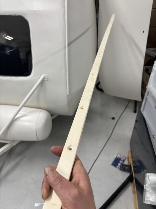 Hand håller en lång trälist med förborrade hål, framför en husvagnsvägg i en verkstadsmiljö.