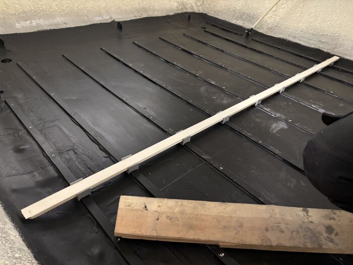 Svart golv i husvagn med tvärgående ribbor av trä, monterade med små stöd. En träbit används som mätverktyg.