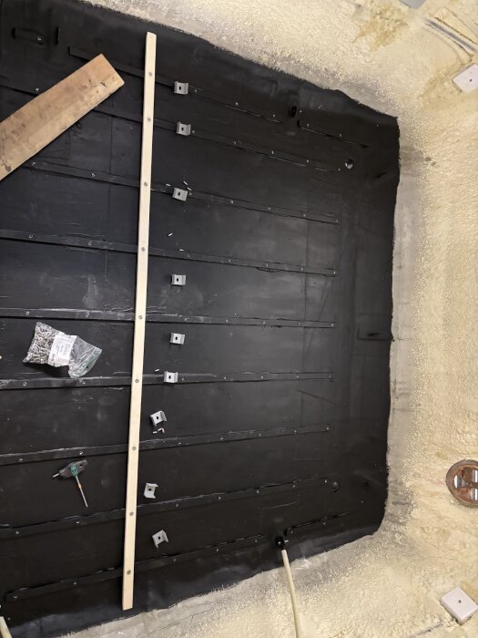 Golvet i husvagn under byggnation, med monterade svartfärgade ribbor, träbit, skruvar och verktyg synliga på ytan.