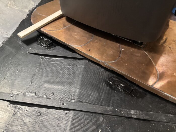 Golvkonstruktion i husvagn med tvärgående ribbor i asp, svart gummimembran och stöd av aluminiumbitar runt aggregatet. Diverse verktyg och material är synliga.