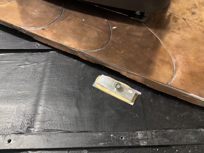 Metallisk styrningsgrej med skruv på svart membran och träskiva i bakgrunden för byggprojekt i husvagn.