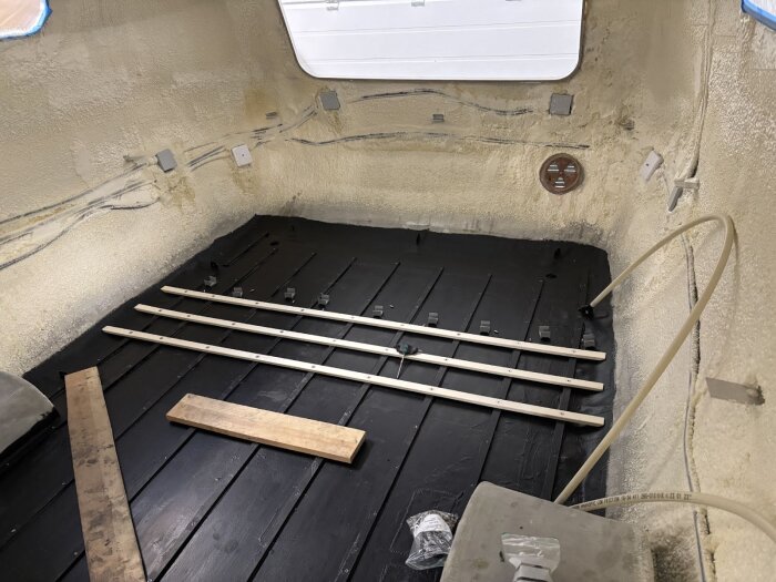 Husvagnsinteriör med nylagd isolering, mörkt golv och tre ljusa träreglar placerade över golvet. Rör och kablar synliga på väggarna.