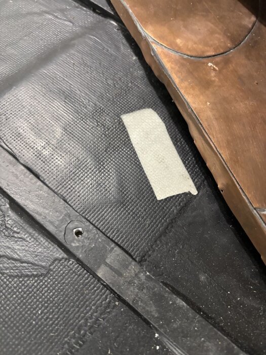 Svart gummimembran med ett hål och tejpbit, bredvid trästruktur i en husvagn under golvkonstruktion.
