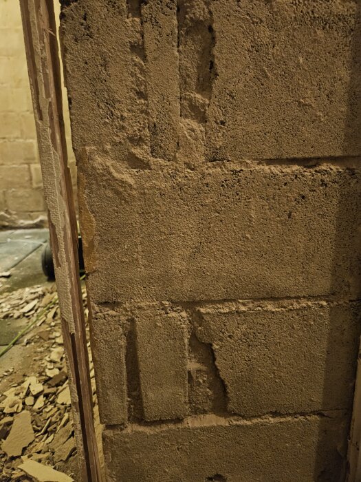 Vägg med synliga betongblock i källaren, där gammal tapet och puts rivits bort. Ytan är ojämn och delvis skadad, redo för renovering.