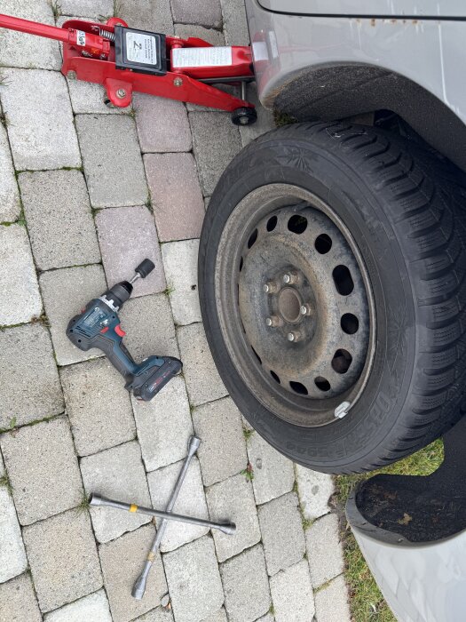 Bil som får däckbyte med domkraft, skruvdragare och fälgkors liggande på stenbelagd uppfart.