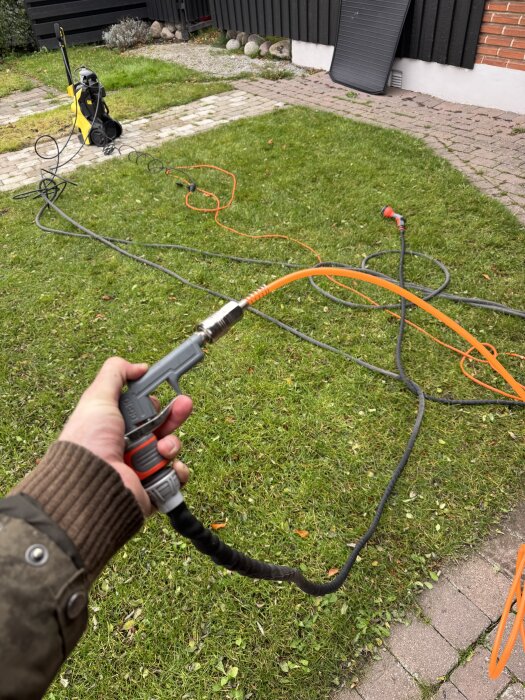 Person blåser rent vattenslang med tryckluft i trädgård, högtryckstvätt synlig i bakgrunden.