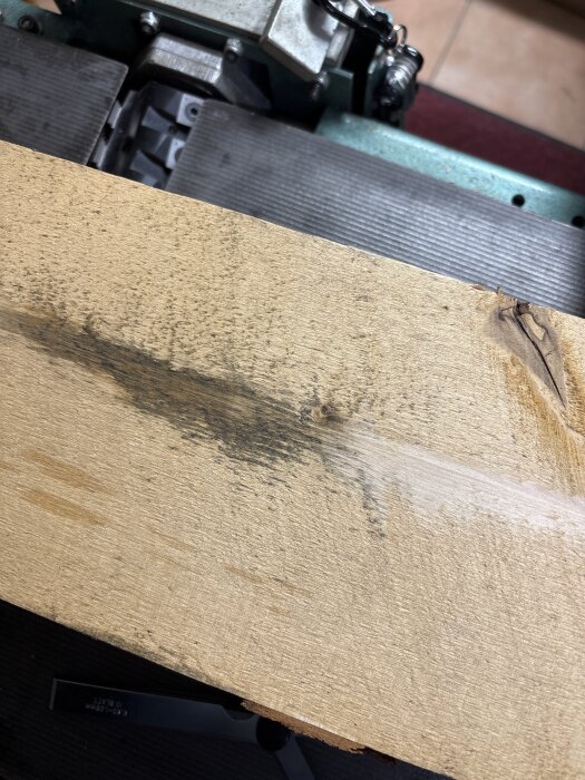 En träbräda med mörka fläckar och verktygsmaskin i bakgrunden, möjligtvis relaterat till behandling av brädans yta.