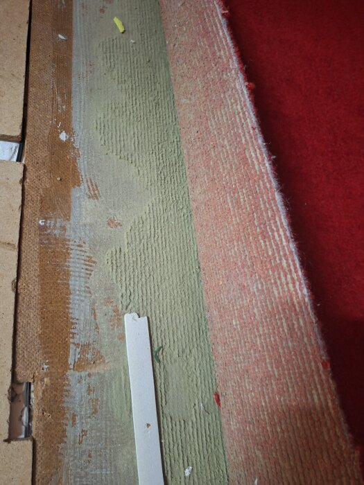 En gammal golvbeläggning under en röd matta, med trä och isoleringsmaterial synligt, från en byggnad uppförd 1973.