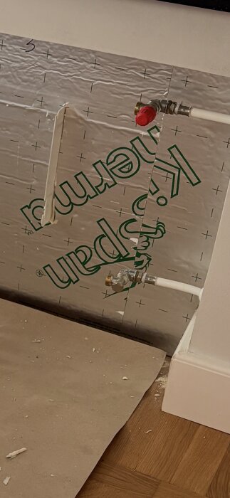 Isolerad vägg med rör och ventiler synliga, golv av trä och papperstäckt yta, skrivbordsskiva delvis synlig.