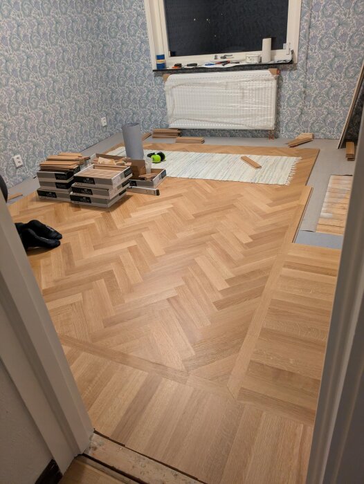 Rum under golvläggning med fiskbensparkett, verktyg och material omkring. Färdigt golv syns i förgrunden, utsmyckat tapet på väggarna.