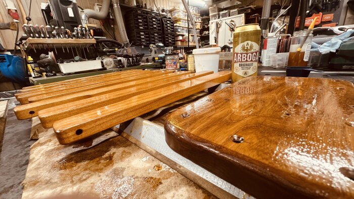 Lackerade trästycken på arbetsbänk med verktyg i bakgrunden, Arboga ölburk bredvid.