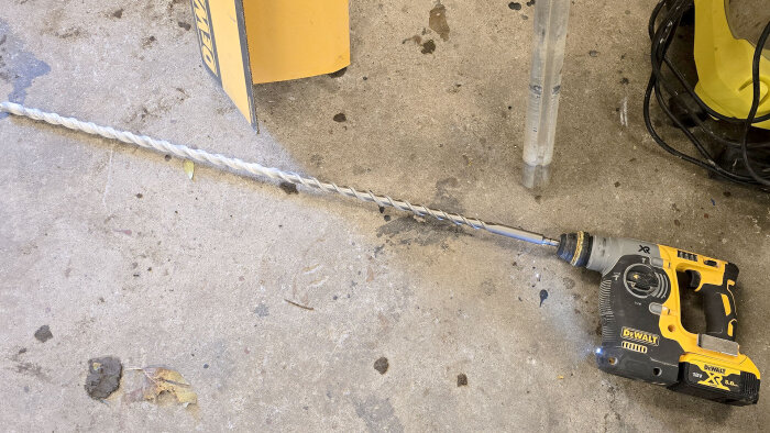 Borrmaskin med lång borr ligger på golvet, bredvid ett verktygsställ och elsladdar i ett garage eller verkstad.