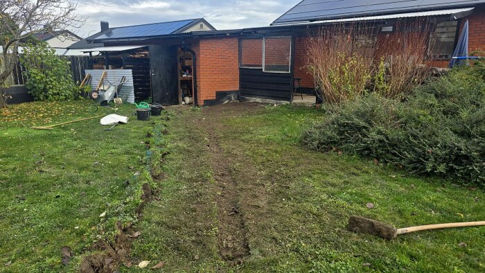 Trädgård med nyligen grävd fåra i gräsmattan framför ett hus med solpaneler på taket. Verktyg och material ligger på gräsmattan.