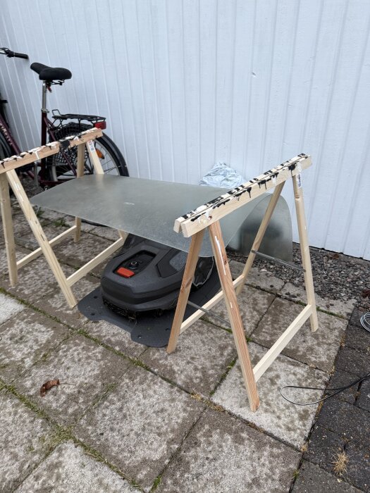 En robotgräsklippare står under ett enkelt tak av träbockar och en platta, intill en vägg med cykel i bakgrunden.