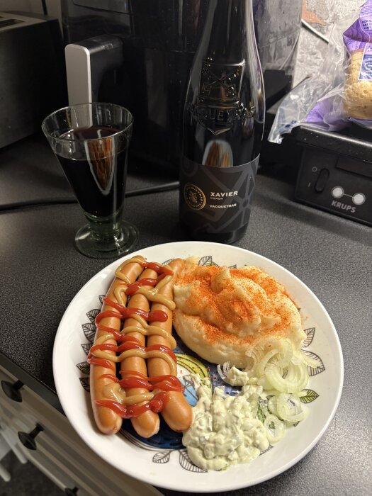 Tallrik med korv och ketchup, potatismos, lök och sås, med ett glas röd dryck och en flaska Xavier Vacqueyras på en bänk.