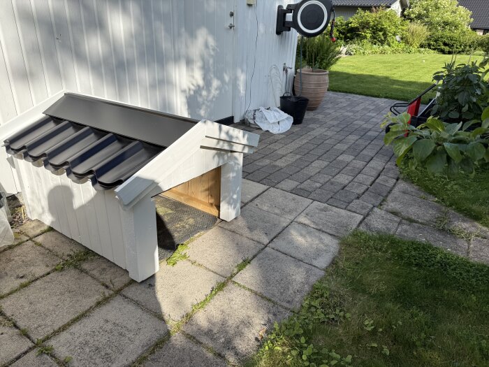 Liten byggnad för robotgräsklippare med svart tak, placerad vid en vit vägg och omgiven av stenplattor och grönska.