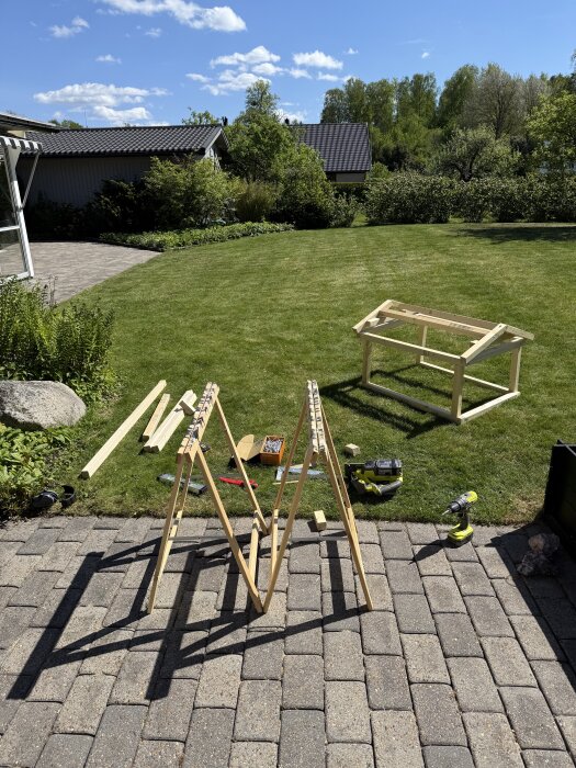 Träram och verktyg på en gräsmatta för att bygga ett hus till en robotgräsklippare, med grönska och hus i bakgrunden.