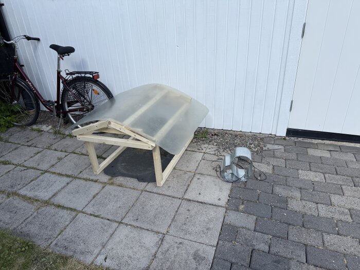 Träplattform med plasttak för robotgräsklippare mot vit vägg, bredvid en cykel.