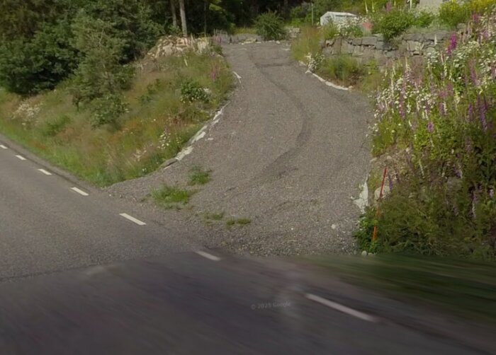 Gruslagd utfart från en 70-väg i innerkurva, omgiven av sten och vildblommor. Anpassad för biltrafik, troligen utan tillstånd.