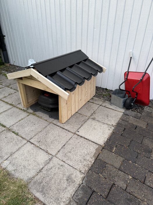 Robotgräsklipparhus med svart tak vid vit husvägg, bredvid en röd skottkärra och stenlagd gång.
