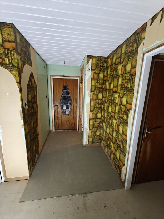 Gammal tapet med grönt och gult mönster delvis borttagen från vägg, med oslipat golv och en stängd dörr i bakgrunden.