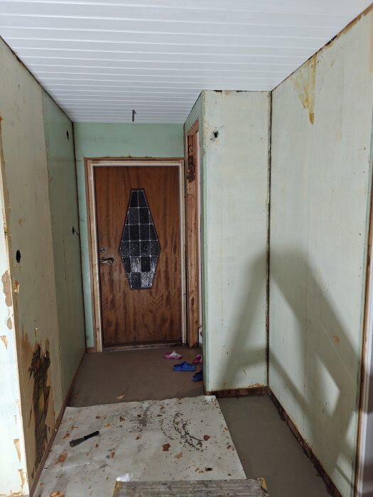 En hall under renovering med bortskrapade tapetrester och lösa skor vid en träytterdörr.