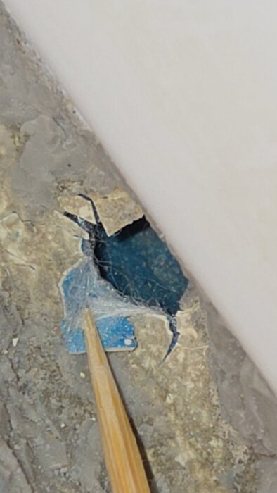 Hål i våtrumsmaterial avslöjar blått och trådigt lager, potentiellt skadat, med en pinne pekande mot det.
