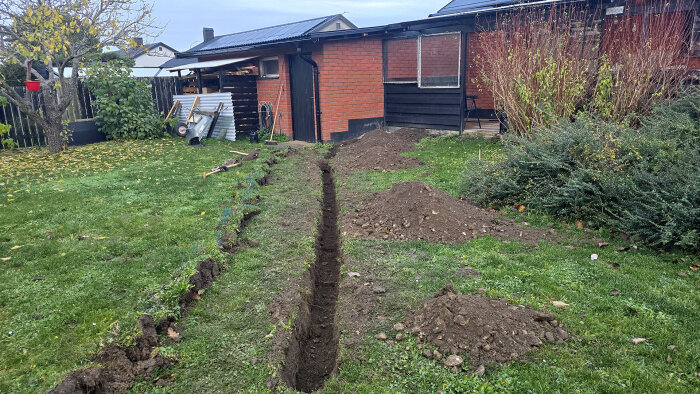 Dike grävt i gräsmatta vid hus, jordhögar runtom, byggmaterial synligt, del av installation för jordtag.