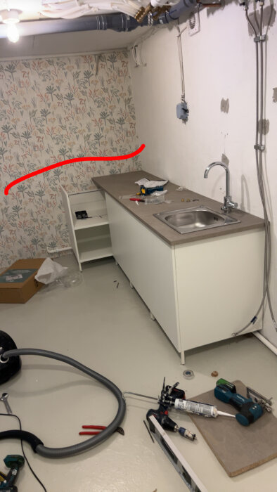 Hörna av tvättstuga med blommigt tapetmönster och en monterad bänkskiva med diskho; verktyg och material i förgrunden.