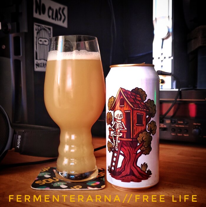 Ett glas ljus öl bredvid en burk med illustration av ett skelett i en trädkoja. Texten "FERMENTERARNA // FREE LIFE" syns på bilden.