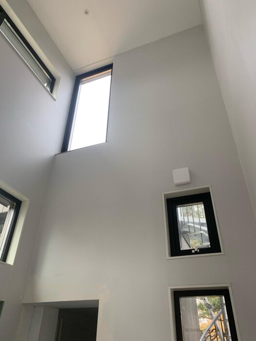 Hög vägg med stora fönsterdörrar, ljusinsläpp ovanifrån. Synligt undertak och del av inomhusmiljö, möjligt byggprojekt.