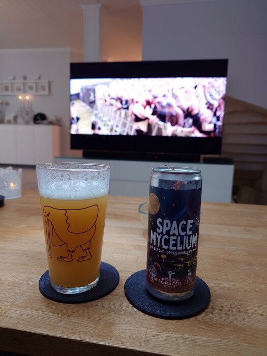 Glas med öl och burk av märket Space Mycelium på ett bord, med en TV i bakgrunden som visar en stor publik.