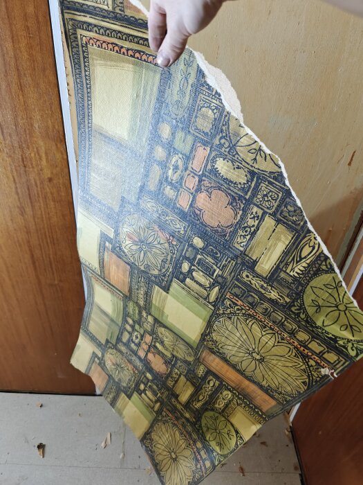 En person håller upp en bit retro tapet med geometriska och blommiga mönster i färgerna grön, orange och gul.