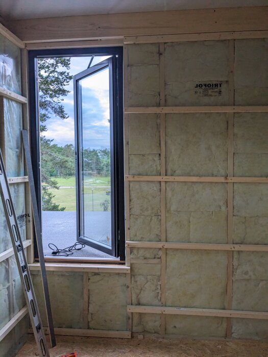 Vägg utan OSB och gips med synlig isolering och träreglar, fönster med utsikt över träd och himmel. En stege står mot väggen.