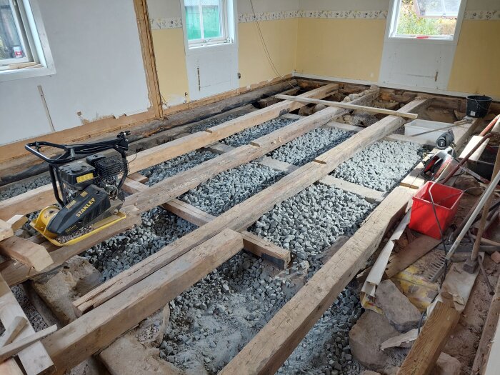 Rum under renovering med bjälklag, krossat grus och verktyg. Utmaning med att förlägga golvvärme mellan de breda bjälkarna.