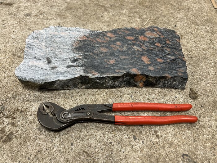 Röd granitprovbit på marken, blöt och glansig, med polygrip nedanför som storleksreferens.