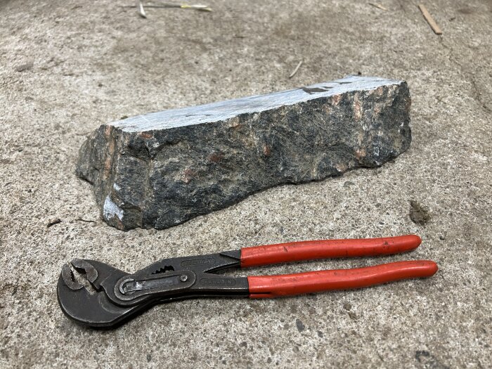 Ett stycke röd granit och en polygrip ligger på en betongyta för storleksjämförelse; graniten har ett rått, ojämnt utseende.