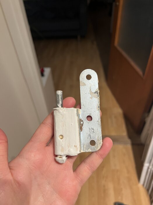 Gammalt vänster gångjärn från Assa Abloy, avskalat och slitet, hålls i hand, del av dörr syns i bakgrunden.