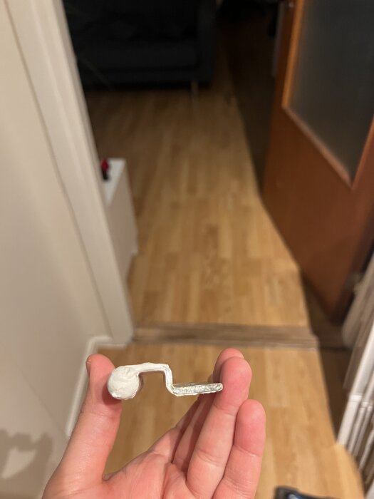 Hand som håller ett vitt vänster Assa Abloy gångjärn från 60-70-talet i ett rum med trägolv och öppen dörr i bakgrunden.