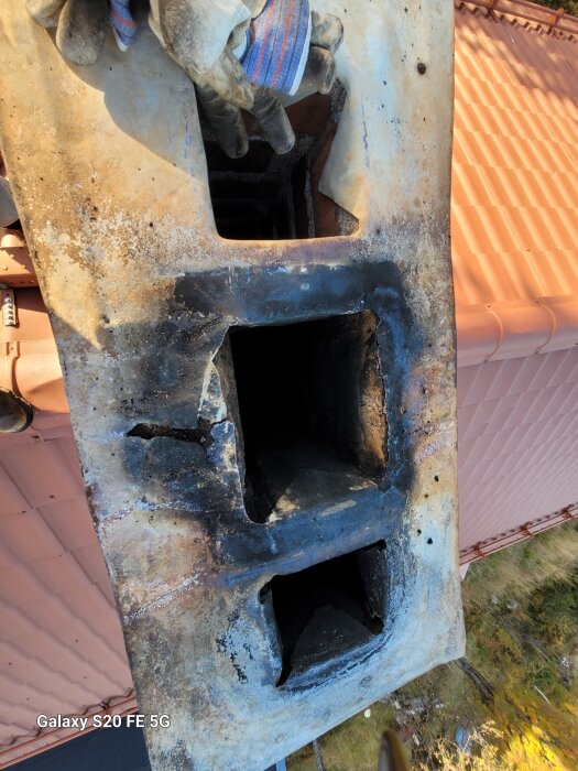 Gammalt skorstenskrön med två rökkanaler synligt ovanför ett tak, omgiven av slitna ytor och handskar i förgrunden.