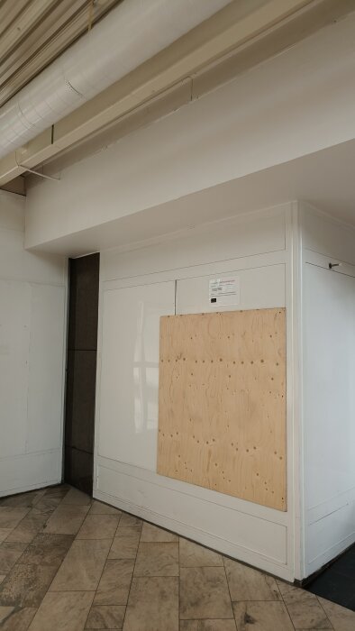 Vägg reparerad med en plywoodskiva och vita paneler, del av en inomhusmiljö med synliga ventilationsrör i taket.