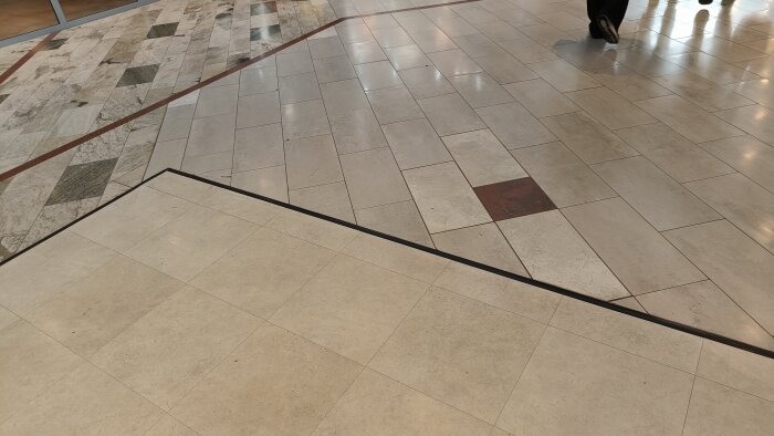 Inomhusgolv med olika plattmönster och en nivåskillnad, del av offentlig byggnad, en person syns i övre högra hörnet.