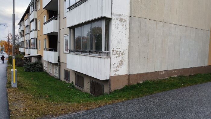 Flerfamiljshus med sliten fasad och balkonger, synliga skador på hörnet av byggnaden; trottoar och grönska i förgrunden.