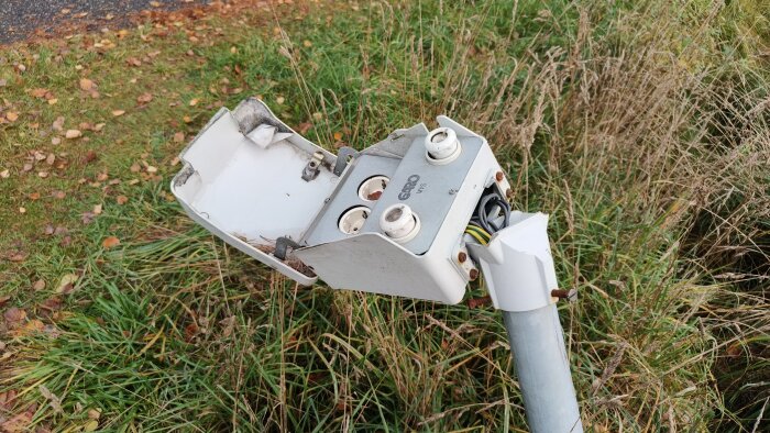 Trasig gatubelysningsarmatur med synliga kablar, placerad i en gräsyta vid en vägkant.