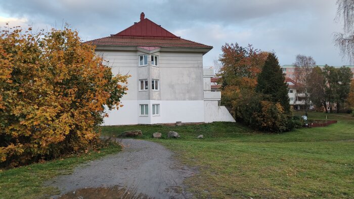 Ett flerbostadshus med rött tak omgiven av höstfärgade träd och en grusväg i förgrunden.