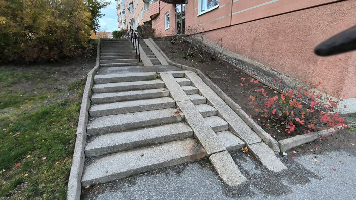 Bred betongtrappa med ojämna steg och ramper vid sidan av ett flerfamiljshus, med buskar och gräsmatta intill.